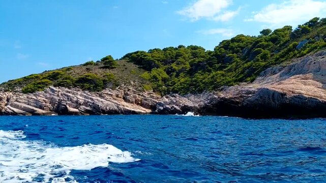 Scenic point-of-view boat journey along the striking rocky coastline of Bisevo Island, Croatia. Lush pine forests top the rugged stone cliffs as deep blue Adriatic Sea waves roll by the vessel