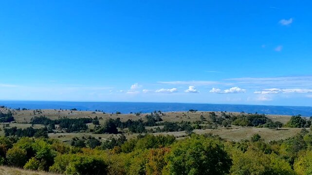 Shot capturing the vast Učka Nature Park, Croatia. A serene, sunny mountain landscape of rolling green hills and scattered trees, with the distant blue Adriatic Sea on the horizon under a clouds