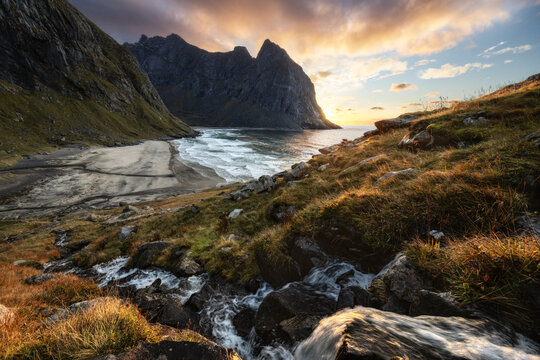 View of Lofoten islands featuring a sandy beach nestled between dramatic mountain peaks and a small stream under a golden sunset sky Vagan Municipality, Nordland, Norway.