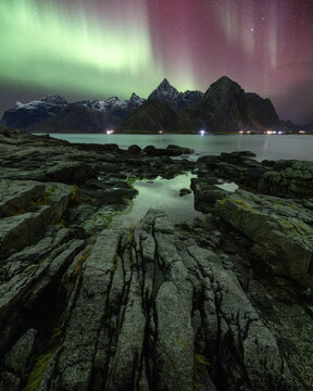 View of the Aurora Borealis dancing over the jagged peaks of Lofoten with rocky coastline and tide pools in Vagan Municipality, Nordland, Norway.