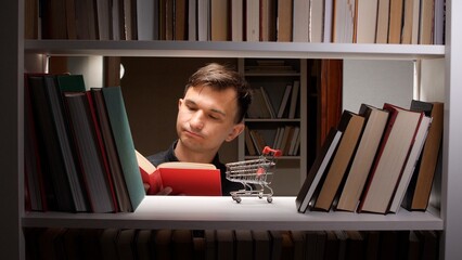 Concentrating man reading a book, standing amongst bookshelves with a miniature shopping cart, symbolizing intellectual consumption and the acquisition of knowledge © Aliaksandr