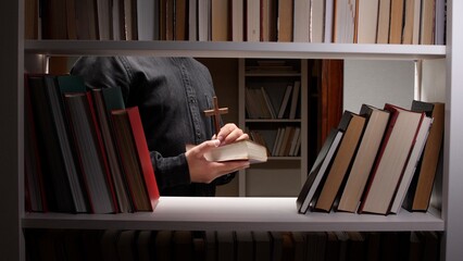 Priest's hands holding a holy bible and a wooden cross, symbolizing faith, religion, and spiritual learning, surrounded by bookshelves filled with religious and theological books © Aliaksandr