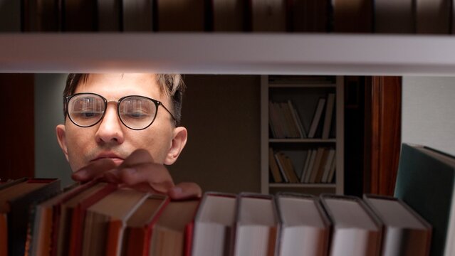 Man wearing glasses looking carefully at books on a library shelf, reaching for one, representing concepts of education, knowledge, academic study, research, and learning pursuit
