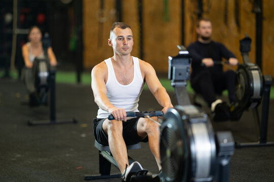 Determined young guy training with rowing machine in sports hall during crossfit workout