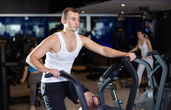 Determined young guy training with air bike in fitness hall during full-body workout