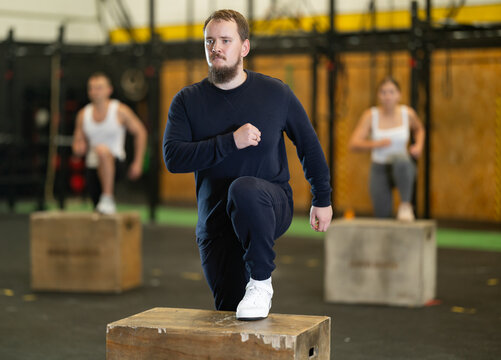 Focused young bearded man in comfortable sportswear participating functional cross training at gym, stepping up onto wooden box to work glutes, quads and hamstrings ..