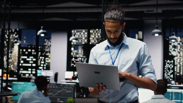 Portrait of smiling middle eastern employee standing in office reading emails on laptop, responding to inbox messages and solving tasks. Man staff analyzing KPIs, interpreting statistical data.