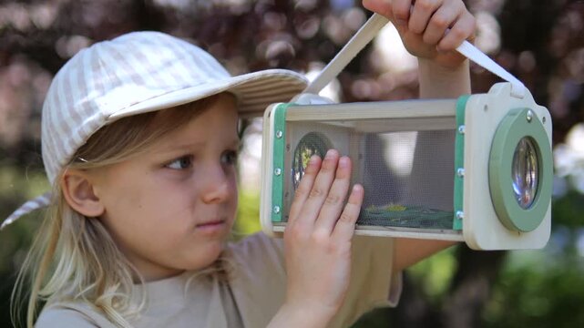 Young explorer kid with a cap closely observing a captured insect inside a wooden bug viewer box, learning about entomology and wildlife in a sunny garden during a summer adventure