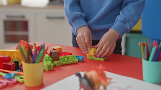 Hispanic boy stacking plastic building blocks with hands on red classroom table surrounded by cups of pencils and art supplies, wearing blue sweatshirt; concentration.
