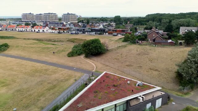 An aerial view showcases a modern triangular building with a green sedum roof situated near a coastal town. Paved paths wind through dry grassy fields leading toward residential houses.