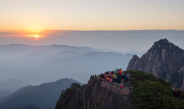 View of a crowd of tourists gathered on a rocky peak of the Huangshan Mountains to watch the sunrise over misty peaks and jagged cliffs in Huangshan City, Anhui, China.
