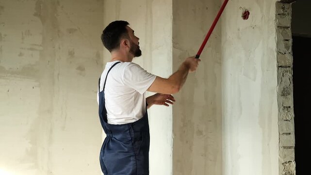 A bearded man in a work uniform is priming the walls after applying gypsum plaster. 