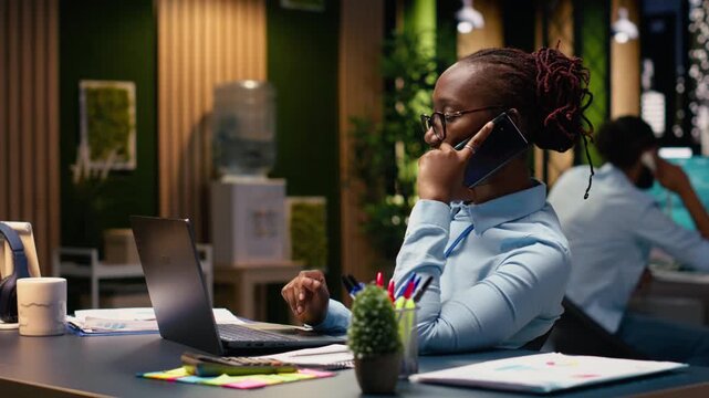 Black employee sharing insights on telephone call in office at night, collaboration between departments to meet objectives and project deadlines. Administrative tasks after hours.
