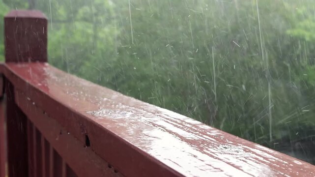 An atmospheric video of a rainstorm. Heavy rain and splashing. Large drops of downpour shatter against the wooden railing of a veranda or balcony. Slow motion.