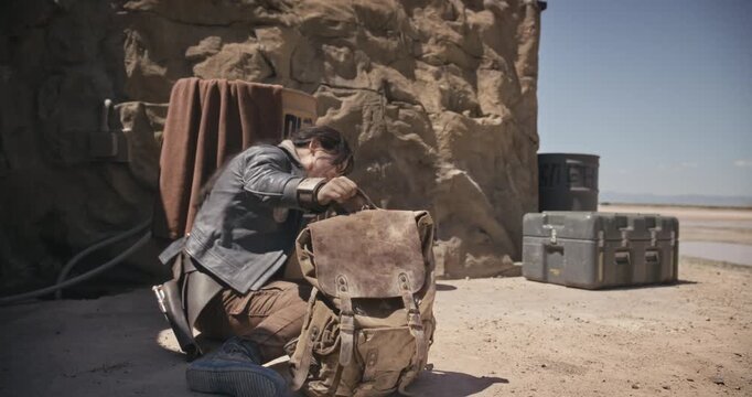 Injured, young, female adventurer resting near barrel with backpack in sci-fi world - wide shot