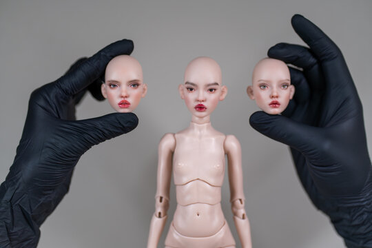 Hands wearing black gloves holding detachable heads amidst doll body. Doll face features unique expressions, highlighting customizable design and craftsmanship of figure