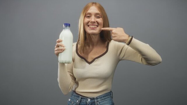 Woman redhead holding milk bottle with left hand and pointing finger to cheek while smiling in studio setting under soft light; wholesome joy.