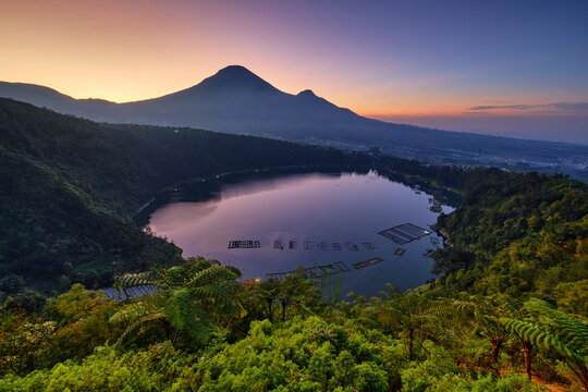 View of Love Valley Seroja Hill overlooking Telaga Menjer lake with fish cages and Mount Sindoro under a colorful sky in Garung, Wonosobo, Central Java, Indonesia.