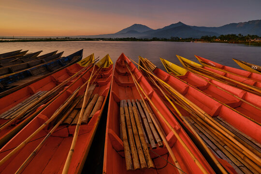 View of colorful wooden boats moored on the calm waters of Rawa Pening lake with mountains under a golden sunset sky in Ambarawa, Central Java, Indonesia.