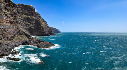 Obraz premium Rocky coastline with dark volcanic rocks and clear blue ocean waves lapping against the shore in a coastal landscape