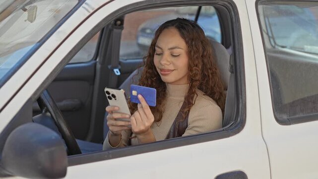 Woman holding smartphone and creditcard, finger pointing to card while seated in a car on a street; digital convenience calm.