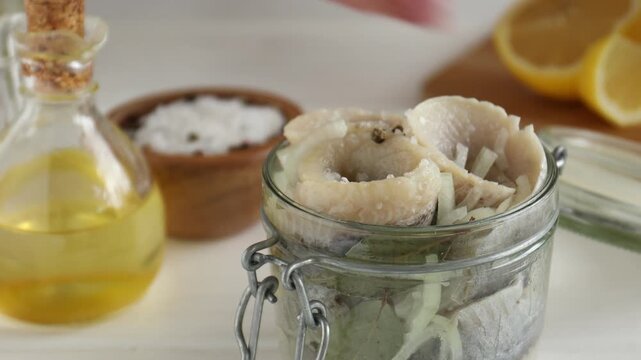 Marinating herring. Woman adding bay leaf, lemon juice and vinegar to fillets in jar at white table, closeup