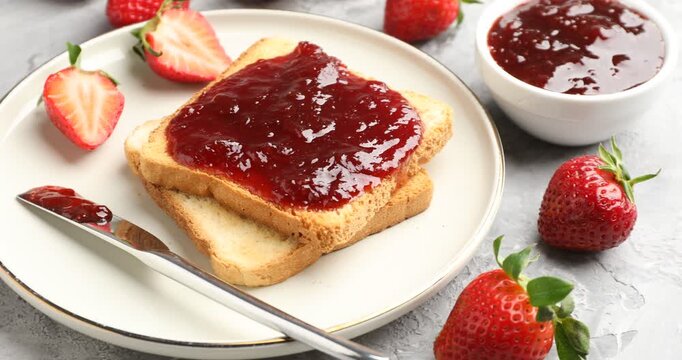Slices of toasted bread with strawberry jam and fresh berries on grey textured table, closeup. Camera moving right