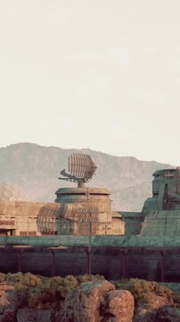 Old buildings stand in a row on a rocky terrain under a clear sky. Large satellite dishes sit atop the structures. Mountains rise in the background. It is daytime and the area looks dry.