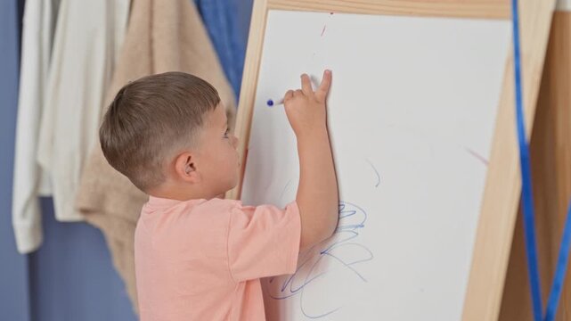 Little hispanic boy drawing on a whiteboard easel with a blue marker, right hand raised, scribbling near a towel and coat in a studio; quiet concentration creativity.