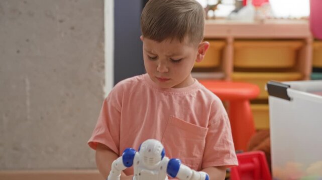 Boy holding a blue and white plastic robot toy and looking to the side showing face and hands, seated near storage bins and a red stool in a building; quiet curiosity.