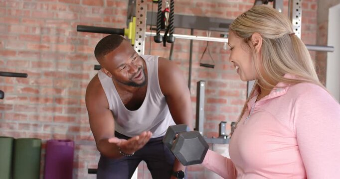 Male trainer leaning and guiding workout-clad female trainee curling hex dumbbell in gym for form