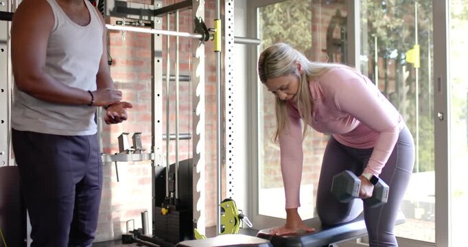 African American trainer giving cues, woman client pulling hex dumbbell on bench, correcting form