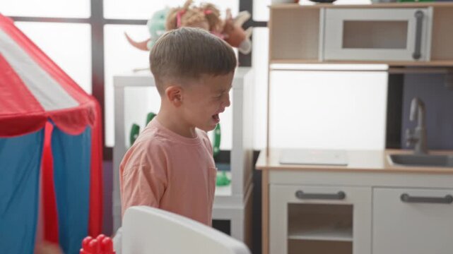 Boy crying beside a play tent and toy dolls, mouth open and hands gesturing, in building; sibling tension sadness.