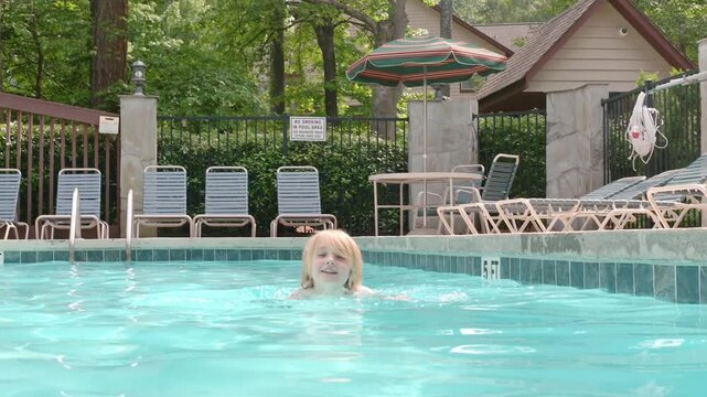 A school-aged child is swimming in a neighborhood pool. A white boy is learning to swim on a hot summer day during the holidays. A blond boy is having fun in an outdoor pool.