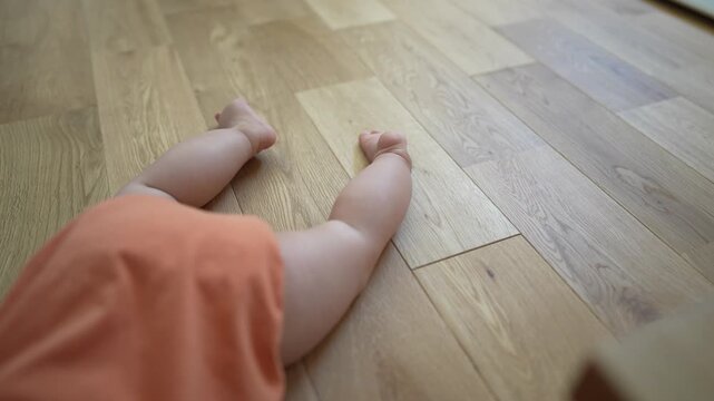 Tiny legs rest on warm wooden floor. Soft orange fabric drapes the baby's body. Calm, curious movements suggest early exploration. Ideal for nursery, parenting, or child development themes