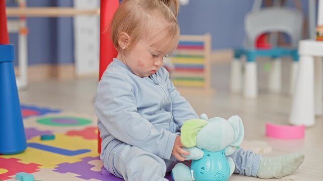 Toddler girl holds a plush elephant toy with small hand while sitting on foam puzzle mat in building; calm curiosity.