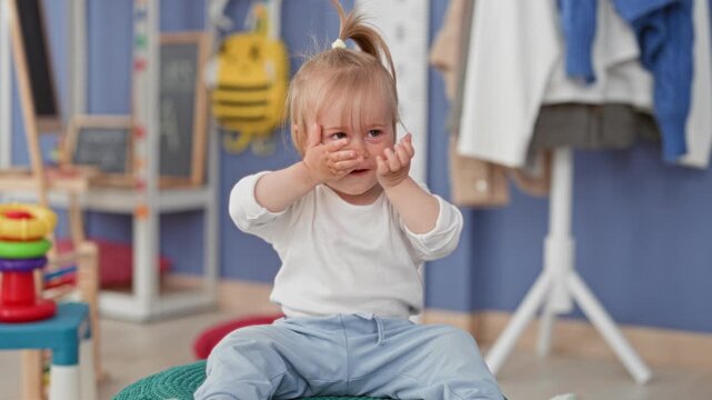 Toddler girl covers her face with both hands in a peekaboo gesture while sitting on a knitted pouf among toys in a playroom; playful curiosity.