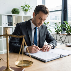 Fototapeta premium Confident male attorney or lawyer signing legal contract documents at desk with scales of justice