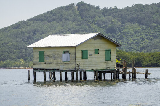 View of a traditional yellow wooden house on stilts over the water with green shutters and lush forested hills in the background Guanaja, Bay Islands Department, Honduras.