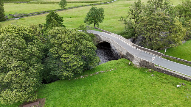 12 July 2025: Aerial view of a stone arch bridge spanning a dark river surrounded by vibrant green pastures, stone walls, and lush trees.