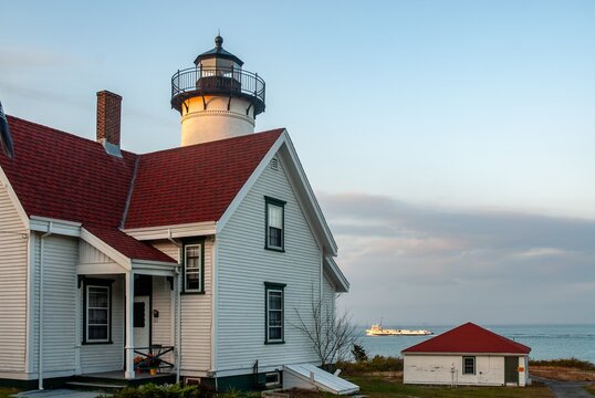 14 May 2026: View of a white lighthouse with a red roof and an attached coastal house with white siding overlooking the sea with a boat in the distance under a soft sky.