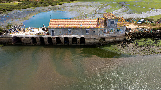 Alhos Vedros, Portugal - 17 December 2024: Aerial view of an old tide mill with stone arches and a terracotta roof situated on the edge of a marshy riverbank.