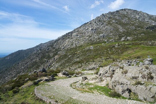 A winding road leads through a rocky landscape with green vegetation and high mountains in the background, old Roman road, at the Puerto del Pico, Sierra de Gredos, &Aacute;vila, Avila, Castilla y Le&oacute;n, Leon, Spain
