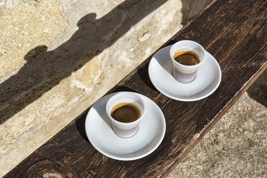 Espresso, Italian coffee speciality in two plastic cups, on a bench made of wood, casting shadows on wall, plastic waste, Sicily, Italy