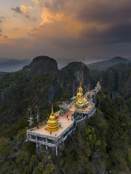 Krabi Noi, Thailand - 22 March 2026: Aerial view of the golden Buddha statue and stupa at the Viewpoint at Tiger Cave Temple atop limestone cliffs during a dramatic sunset.