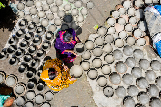 Munshiganj, Bangladesh - 28 September 2021: Aerial view of two women in colorful saris arranging handmade clay pots on the ground to dry in the sun.