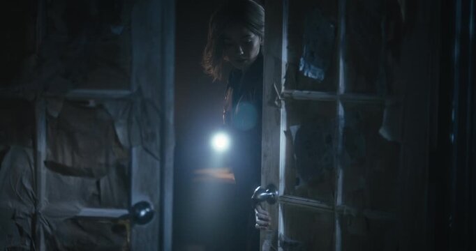 Young woman with flashlight exploring scary, dark room then propping door open with book - steady cam shot