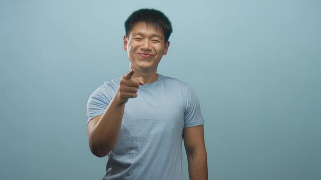 Man pointing finger at camera with confident grin, wearing light blue t shirt, bare forearm visible, studio lighting and seamless backdrop; playful approval.