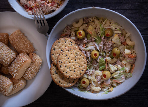 Ensalada de atun con lechuga mayonesa y aceitunas rellenas, con galletas de ajonjoli encima dentro de un tazon blanco, vista cenitall con bollitos al lado