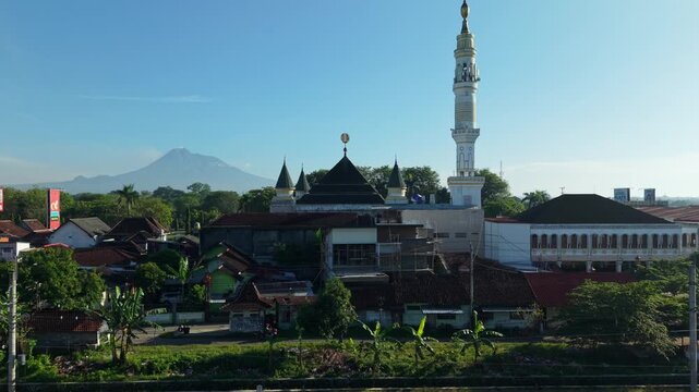 Aerial view of a mosque with a tall minaret and Mount Merapi in the background with railway tracks and greenery in Kecamatan Prambanan, Daerah Istimewa Yogya, Indonesia.